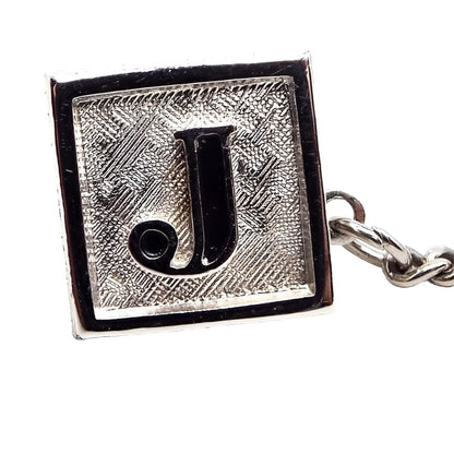 Enlarged front view of the Mid Century vintage Swank initial tie tack. It's square in shape and has matte brushed textured silver tone on the front. There is a block style letter J in the middle that is black enameled on the inside.