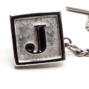 Enlarged front view of the Mid Century vintage Swank initial tie tack. It's square in shape and has matte brushed textured silver tone on the front. There is a block style letter J in the middle that is black enameled on the inside.