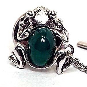 Close up view of the Mid Century vintage jelly belly frog tie tack. The metal is darkened silver tone in color. It is a view of the top side of the frog from above. There is an oval dark green glass cab in the middle for the body.