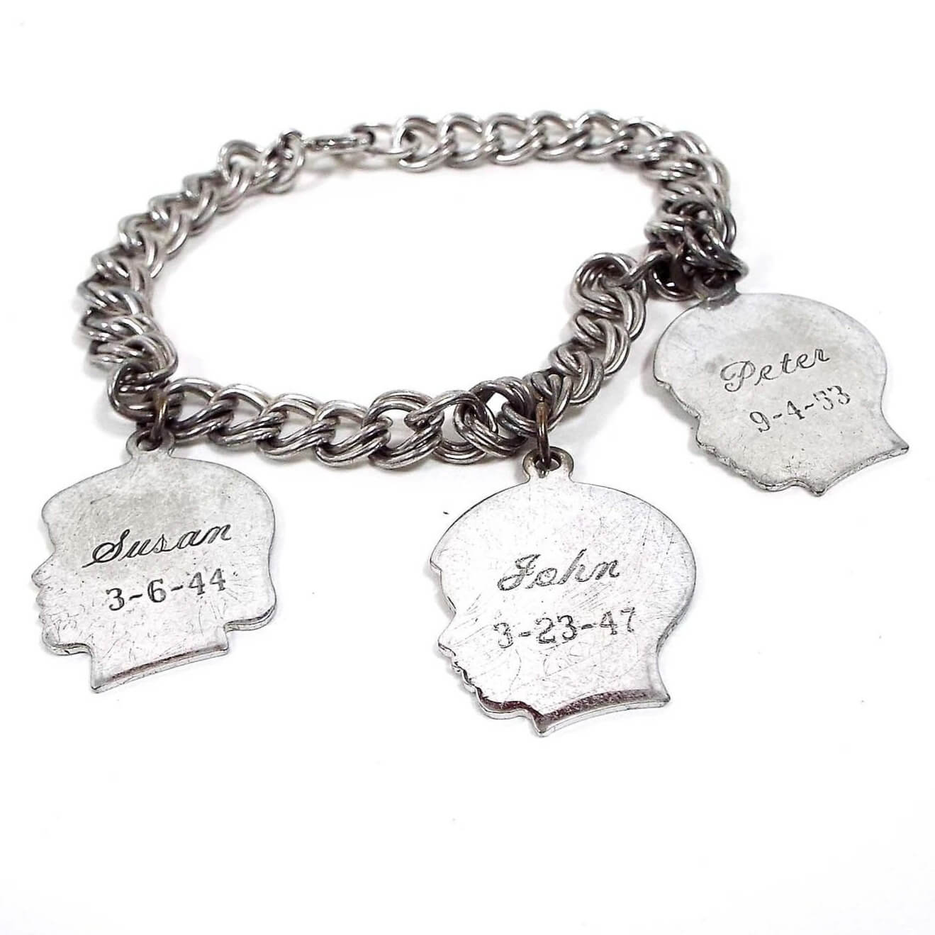 Front view of the Mid Century vintage sterling silver charm bracelet. It is silver tone in color with double link curb chain. There are three head shaped charms. Two are boy shaped and one girl. They have names and birthdates of Susan 3-6-44, John 3-23-47, and Peter 9-4-53. 