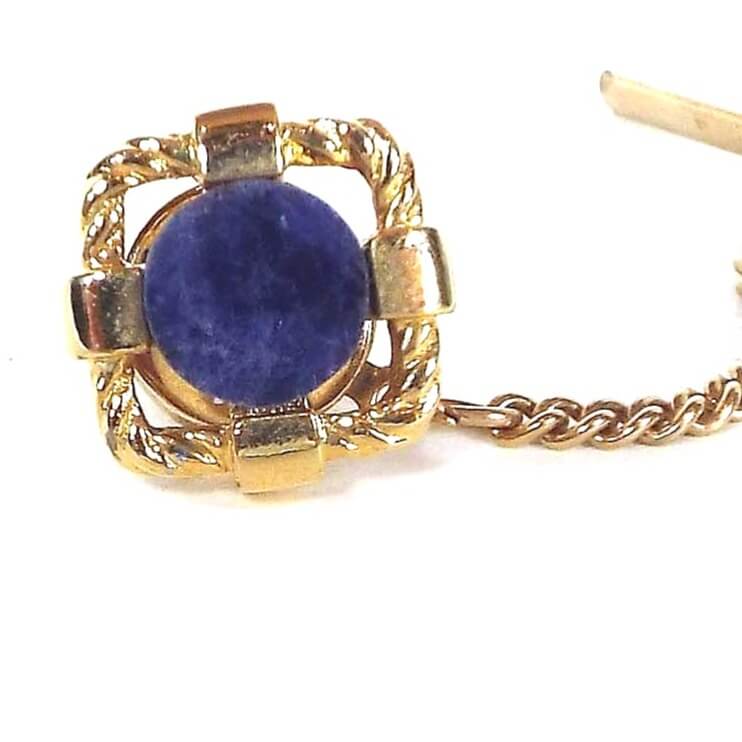 Enlarged front view of the retro vintage gemstone tie tack. It has a rounded square shape with an open middle. In the middle is a round flat sodalite gemstone cab in denim blue color. The metal is gold tone in color.