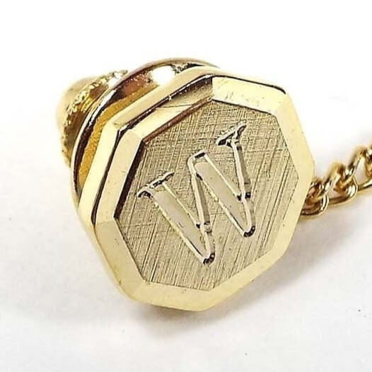 Front view of the Mid Century vintage initial tie tack. It is gold tone in color and has a faceted octagon shape. The front is textured and there is an engraved letter W in the middle.