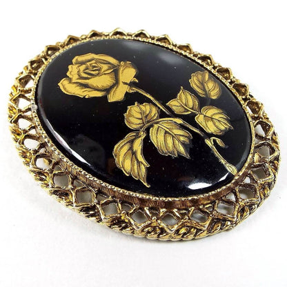 Angled front view of the Mid Century vintage brooch pin. It's antiqued brass in color with an filigree oval setting. The middle has a black glass cab with a metallic gold painted rose flower on it.