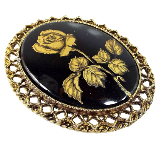 Angled front view of the Mid Century vintage brooch pin. It's antiqued brass in color with an filigree oval setting. The middle has a black glass cab with a metallic gold painted rose flower on it.