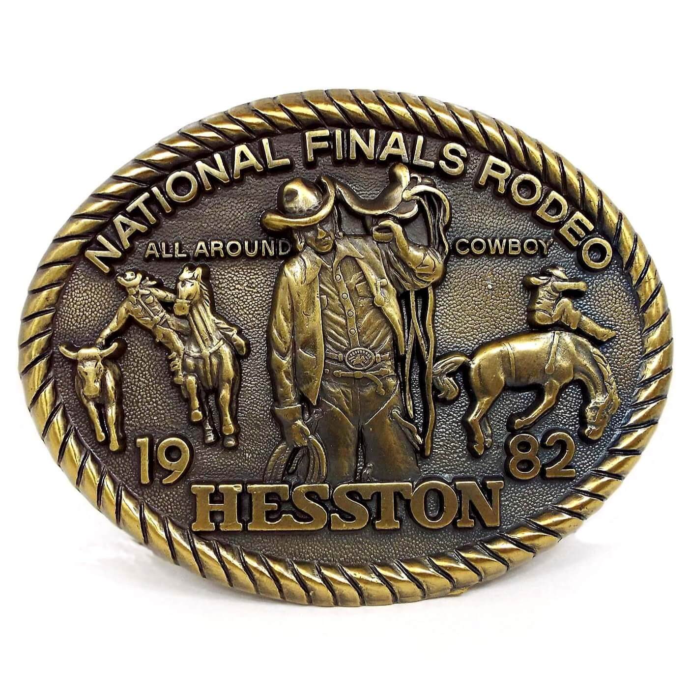 Front view of the retro vintage Hesston 1982 belt buckle. It is oval and antiqued brass in color. It says National Finals Rodeo All Around Cowboy 1982 Hesston on the front. There are depictions of a cowboy roping cattle, riding a bucking bronco, and carrying a horse saddle.