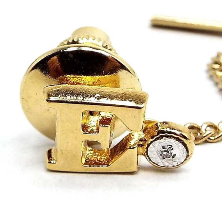 Enlarged front view of the retro vintage initial tie tack with diamond chip. The metal is gold tone plated in color. The tie tack is shaped like a block letter E. To the right of the letter is a small round area with a tiny diamond chip in it surrounded by faceted silver tone metal.