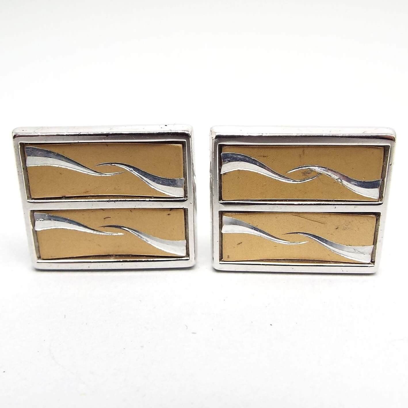 Front view of the retro vintage Anson two tone cufflinks. They are square in shape with the front being divided into two rectangle areas with wavy spikes pointing towards the middle. The metal is mostly silver tone in color with an antiqued gold tone color background. 