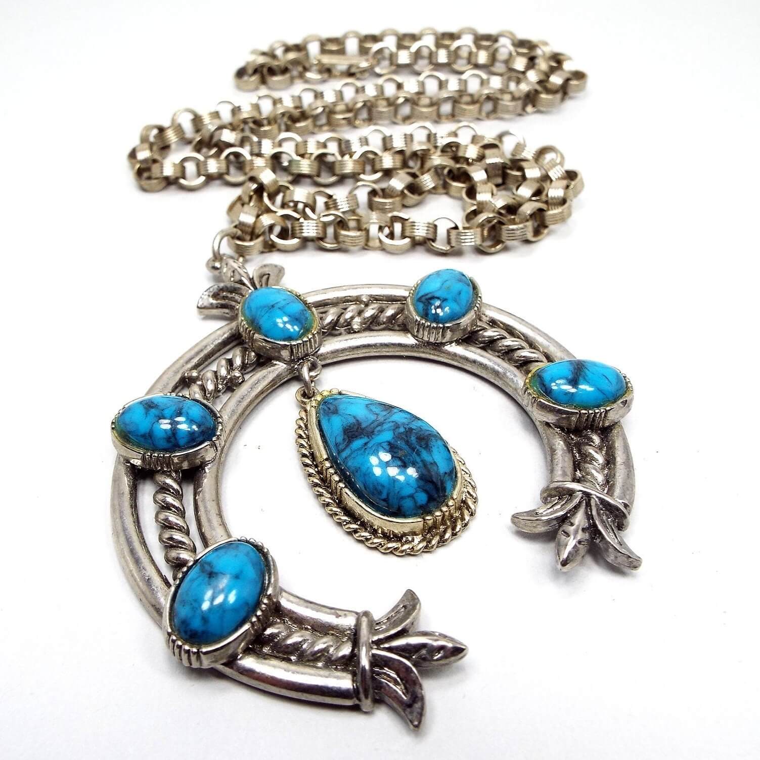 Front view of the retro vintage ART squash blossom pendant necklace. The metal is silver tone in color. The pendant has a large curved squash blossom design with oval blue and gray marbled plastic cabs. 