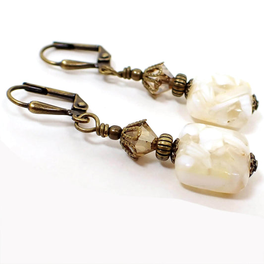 Angled view of the handmade square drop earrings. The metal is antiqued brass in color. There are faceted glass crystal beads at the top in a light golden brown. The bottom beads are off white resin squares with pieces of white and off white mother of pearl shell.