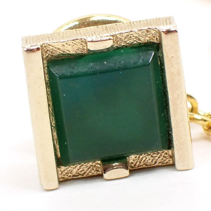 Enlarged front view of the Mid Century vintage Swank tie tack. It is gold tone plated in color and has a square shape. There is a square green glass faux jade cab on the front.