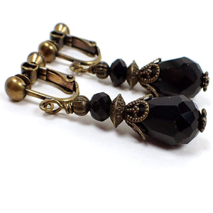 Angled side view of the handmade vintage style black teardrop earrings. The metal is antiqued brass in color. There is a faceted glass rondelle black bead at the top and a faceted teardrop black bead at the bottom. 