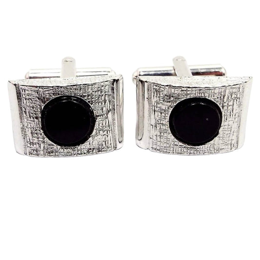 Front view of the retro vintage silver tone and black cufflinks. The have a domed rectangle shape on the front with a flat round black glass cab in the middle. The silver tone metal on the front is textured.