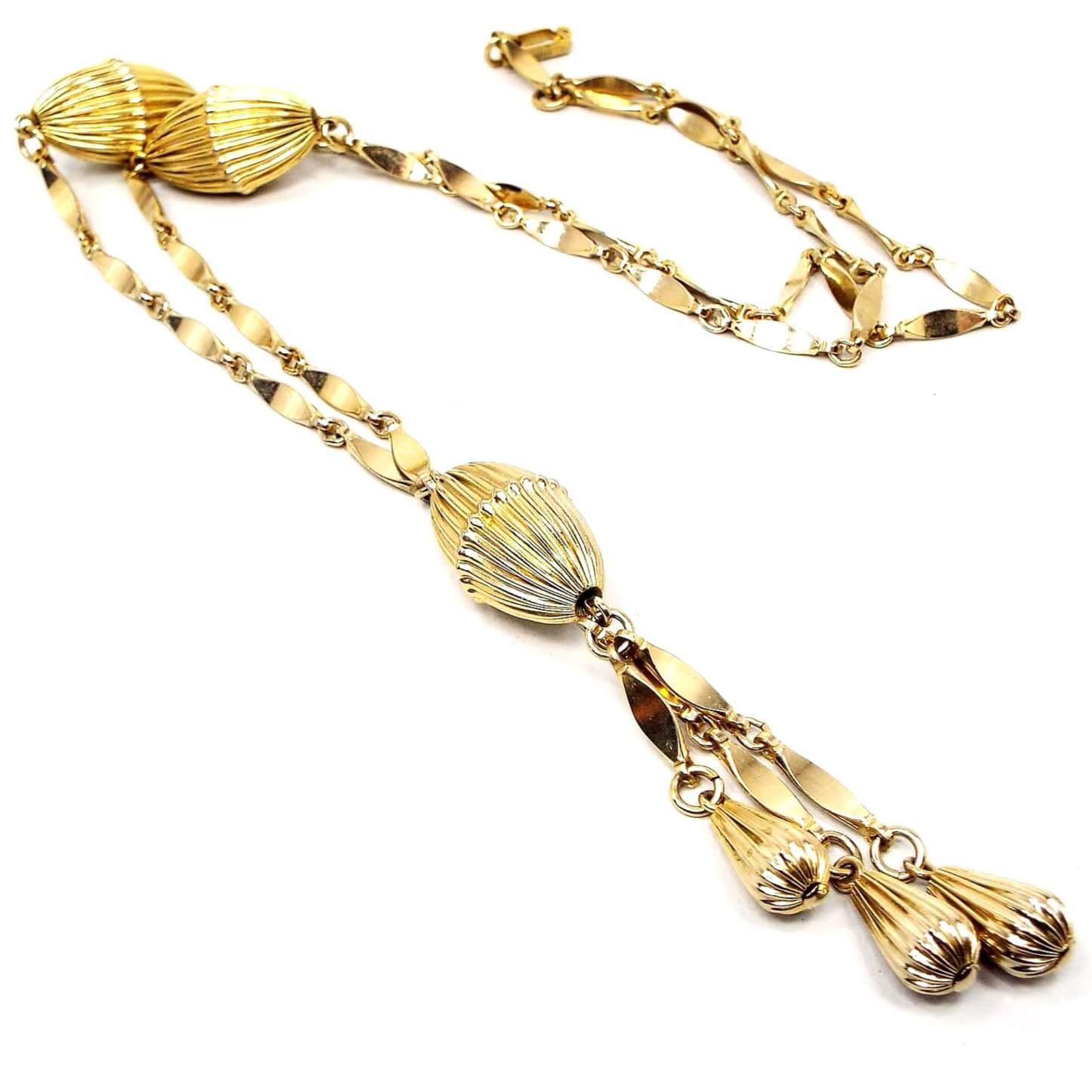 Angled view of the Mid Century vintage tassel necklace. The metal is gold tone in color. There are marquis shaped links down to two large oval corrugated beads and then more links to the bottom tassel. The tassel area has one large oval corrugated bead and then three strands of marquis link chain down to smaller sized corrugated teardrop shaped beads at the bottom.