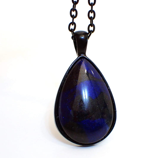 Enlarged front view of the handmade Goth resin pendant. The metal is black coated. The pendant is teardrop shaped with marbled pearly black and cobalt blue color resin.
