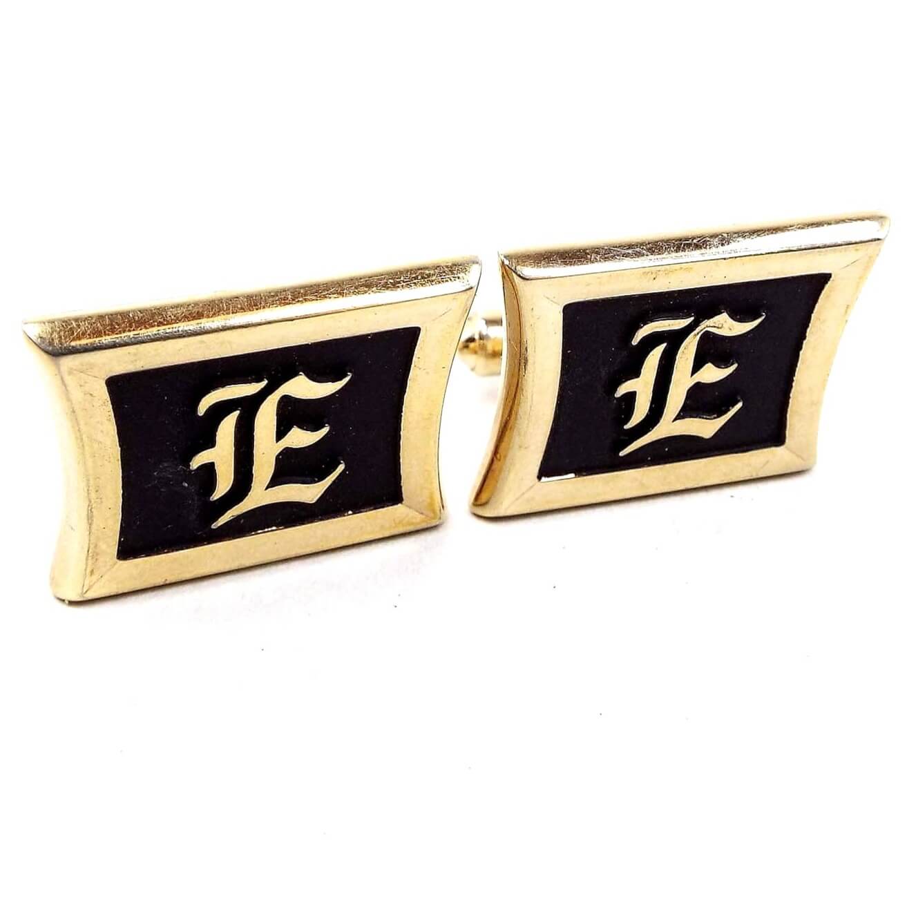 Front view of the Mid Century vintage Hickok initial cufflinks. They are are rectangle in shape with pinched sides. The metal is gold tone in color. The fronts are black painted with a fancy gold tone color letter E in the middle.