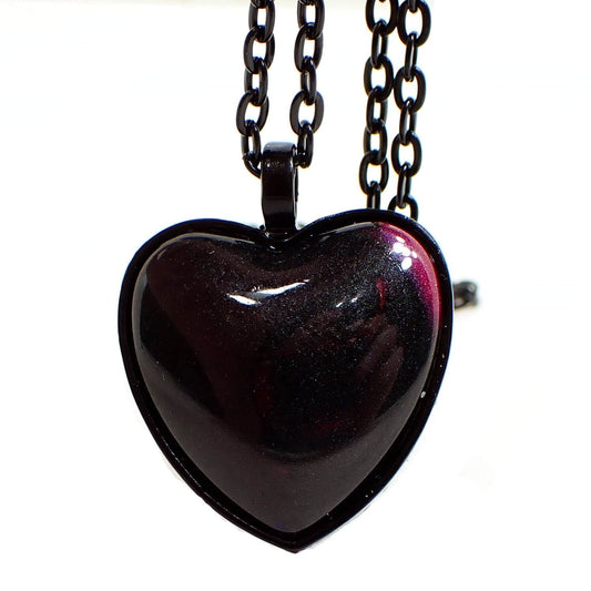 Front view of the handmade resin black heart Goth pendant necklace. The chain and pendant setting are black coated. The domed resin heart cab is mostly shimmery black in color with a hint of bright pearly pink at the corner.