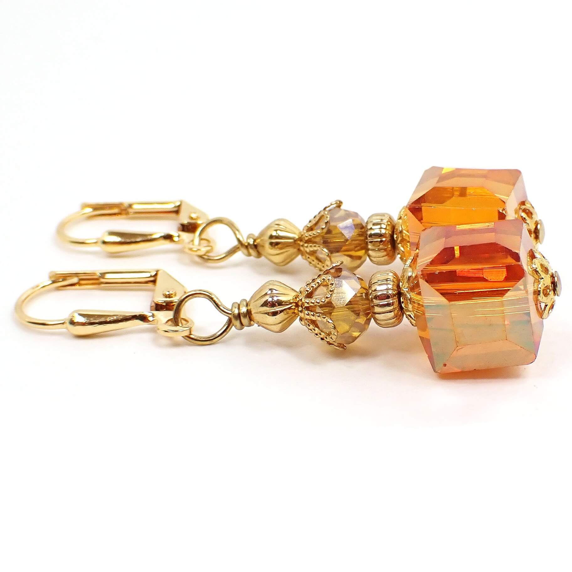 Side view of the handmade cube drop earrings. The metal is gold plated in color. There are faceted oval glass crystal beads at the top in a light orange color. The glass crystal cube beads at the bottom are mostly orange with hints of pink and yellow as you move around in the light.