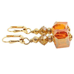 Side view of the handmade cube drop earrings. The metal is gold plated in color. There are faceted oval glass crystal beads at the top in a light orange color. The glass crystal cube beads at the bottom are mostly orange with hints of pink and yellow as you move around in the light.