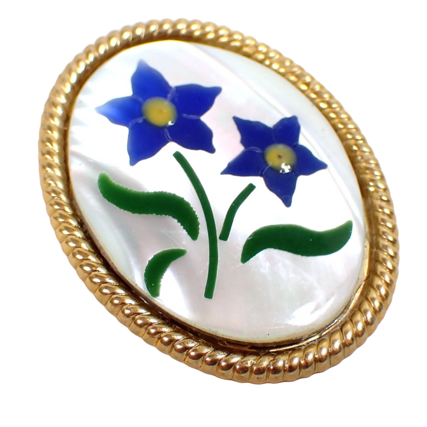 Angled front view of the retro vintage floral brooch pin. It is a large oval shape with gold tone plated metal edge. There is a pearly white mother of pearl cab on the front. There are pieces of dyed shell inlaid in the mother of pearl cab to form two blue flowers with yellow centers and green leaves.