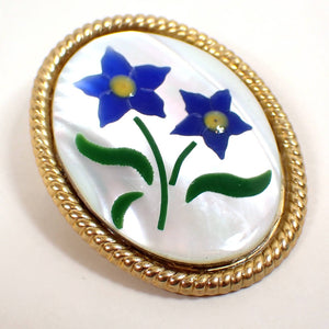 Angled front view of the retro vintage floral brooch pin. It is a large oval shape with gold tone plated metal edge. There is a pearly white mother of pearl cab on the front. There are pieces of dyed shell inlaid in the mother of pearl cab to form two blue flowers with yellow centers and green leaves.
