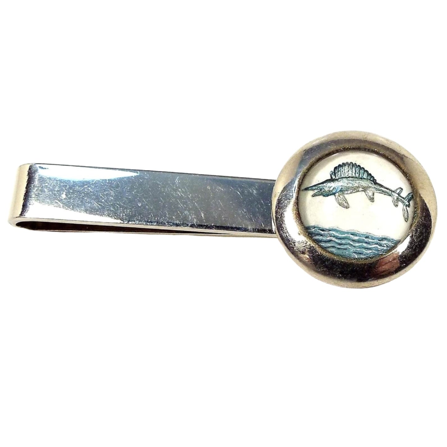 Front view of the Mid Century vintage sailfish tie bar. It is a very light gold tone in color. There is a round glass cab at the end with a design of a sailfish or marlin jumping out of the water over the waves. 