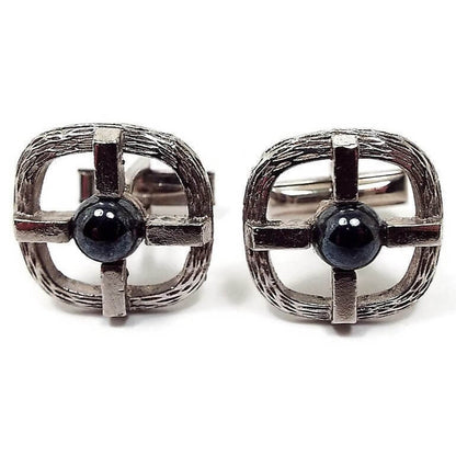 Front view of the retro vintage imitation hematite cufflinks. They have a textured silver tone rounded square design that's open in the middle. Over that is a silver tone metal cross that has a domed round faux hematite cab in the middle.