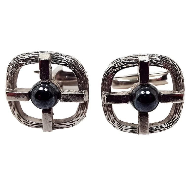 Front view of the retro vintage imitation hematite cufflinks. They have a textured silver tone rounded square design that's open in the middle. Over that is a silver tone metal cross that has a domed round faux hematite cab in the middle.