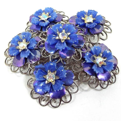 Front view of the Coro Mid Century vintage rhinestone floral brooch pin. The metal is a slightly darker silver tone in color. There are five blue flowers around another in the middle of the brooch. Each flower has jagged edge blue plastic petals on top with rounded AB blue enameled metal petals underneath. There are AB clear rhinestones in the middle of each flower.