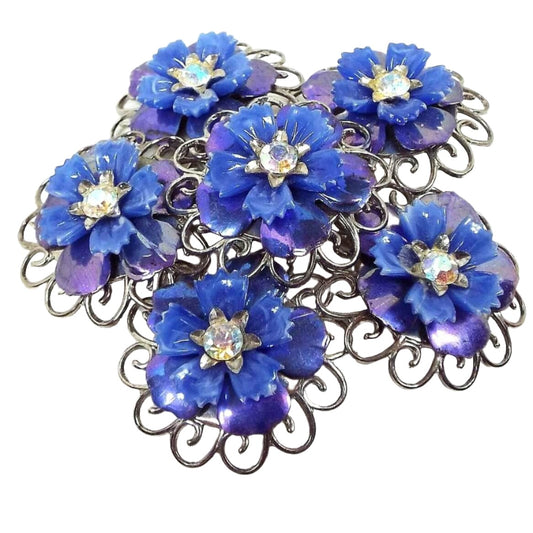 Front view of the Coro Mid Century vintage rhinestone floral brooch pin. The metal is a slightly darker silver tone in color. There are five blue flowers around another in the middle of the brooch. Each flower has jagged edge blue plastic petals on top with rounded AB blue enameled metal petals underneath. There are AB clear rhinestones in the middle of each flower.