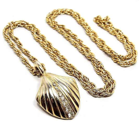 Front view of the retro vintage TAT shell shaped rhinestone pendant necklace. The metal is gold tone color. There is a rope style chain down to a shell shaped pendant with grooves. The middle groove has a row of clear round rhinestones going down it. The rhinestones get larger as it goes down the pendant.