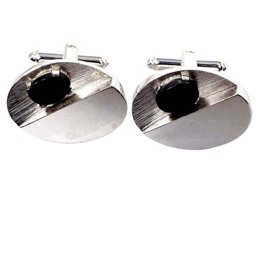 Front view of the Dante Mid Century vintage cufflinks. They are silver tone in color and oval in shape. On one half of the oval there is an indent with a prong set oval black glass cab on it. 