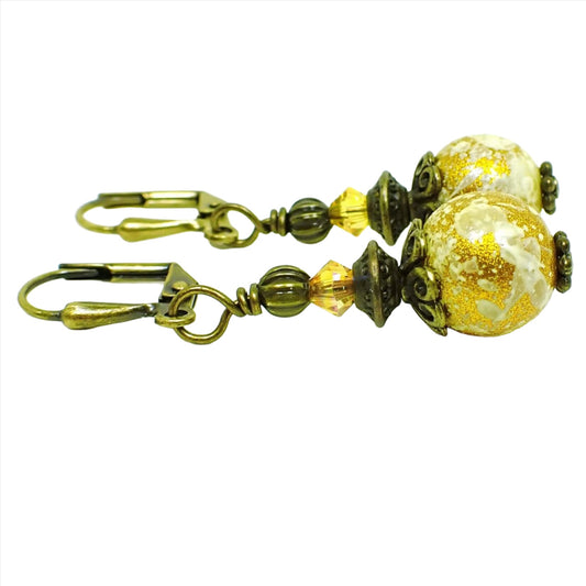 Side view of the handmade glass drop earrings. The metal is antiqued brass in color. There is a faceted glass crystal bead at the top in a bright yellow color. The bottom round glass bead is off white with splashes of metallic gold color.