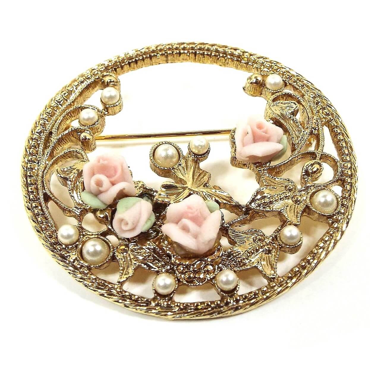 Front view of the Mid Century vintage faux pearl and floral brooch pin. The metal is gold tone in color. The brooch is round has a a floral design with vines around the sides and bottom. There are plastic faux pearls scattered here and there around the edge. There are four light pink porcelain roses with green leaves at the bottom area.