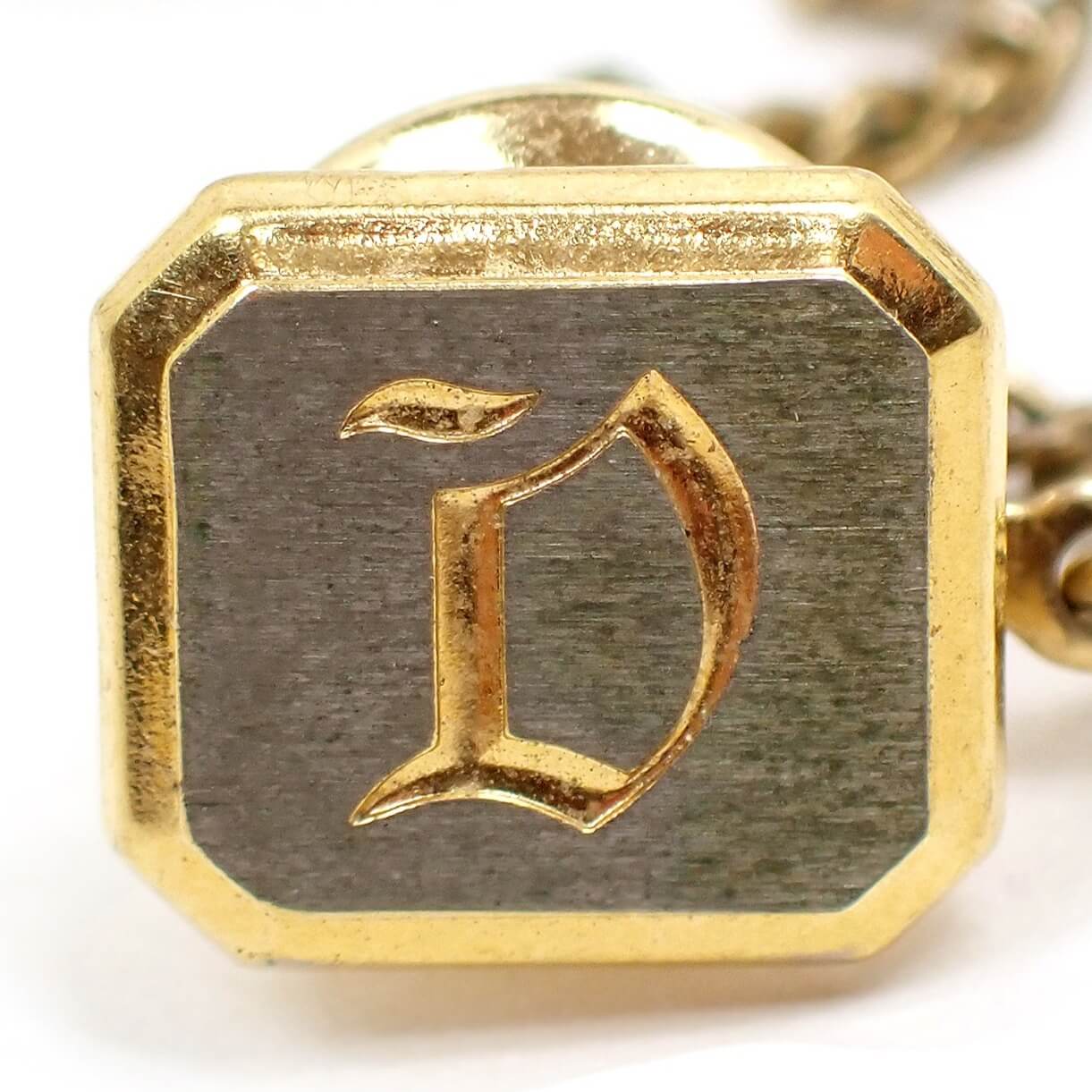 Enlarged front view of the Mid Century vintage initial tie tack. It is octagon shaped with brushed matte silver tone front. The edge and clutch are gold tone plated in color. There is a gold tone fancy style block letter D engraved on the front.