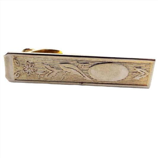 Front view of the Mid Century vintage floral tie clip. It is a darkened gold tone in color. The are stripes inside the rectangle with a raised flower design, some leaves, and an oval on the end. 