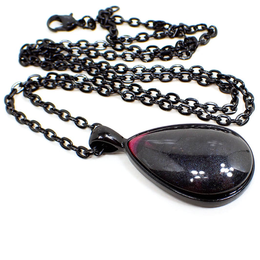 Angled view of the handmade resin teardrop pendant necklace. The chain and setting are black in color. The pendant is teardrop shaped with a domed resin cab that has pearly black resin with a touch of bright pink at the top. 