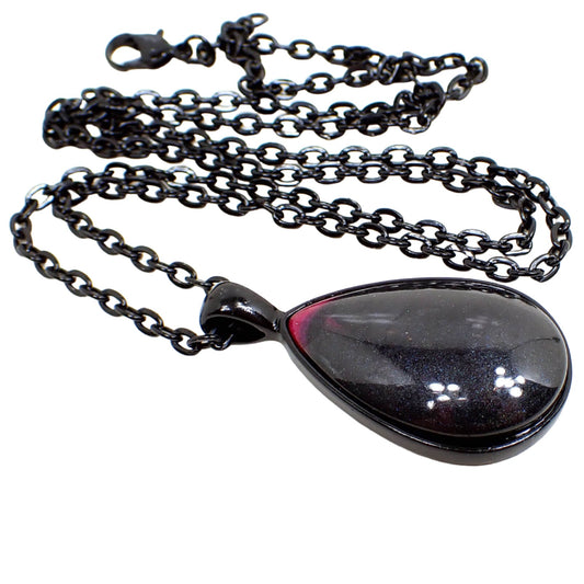 Angled view of the handmade resin teardrop pendant necklace. The chain and setting are black in color. The pendant is teardrop shaped with a domed resin cab that has pearly black resin with a touch of bright pink at the top. 