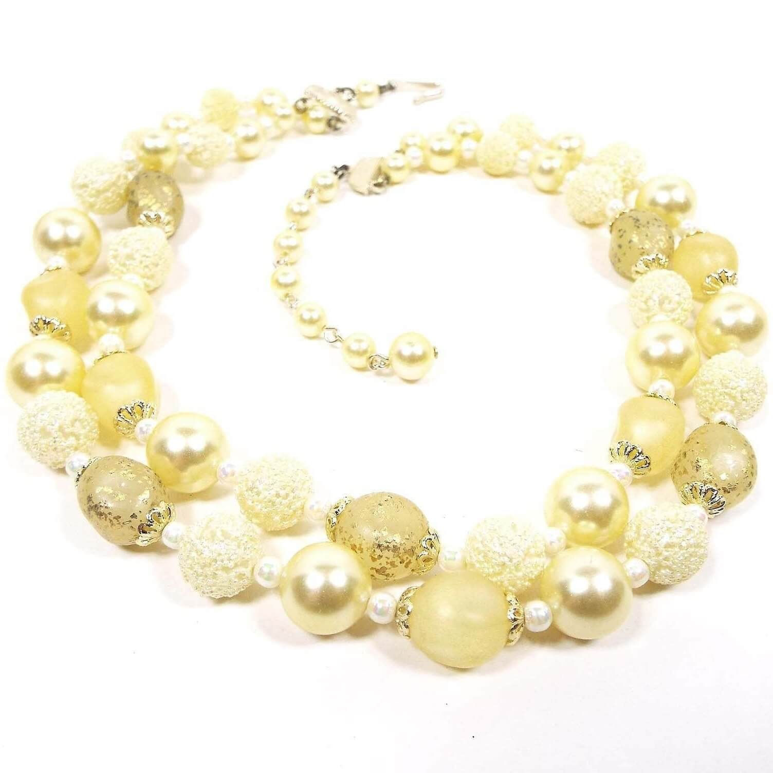 Angled view of the Mid Century vintage multi strand necklace from Japan. The beads are varying shades of light yellow. There are pearly beads, metallic gold speckled beads, nugget shape beads, and bumpy textured "sugar" beads. There is a hook clasp at the end.