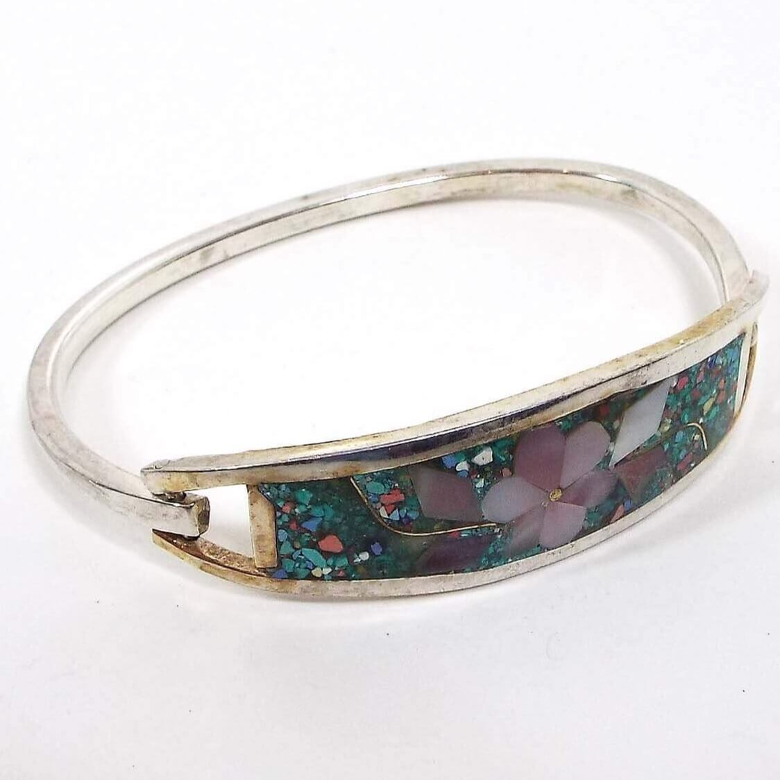 Angled view of the retro vintage Taxco hinged bangle bracelet. The metal is slightly darkened silver tone color from age. The front part has a dyed pink inlaid mother of pearl shell flower design. The rest of the front is filled in with teal green resin and multi color stone chips.