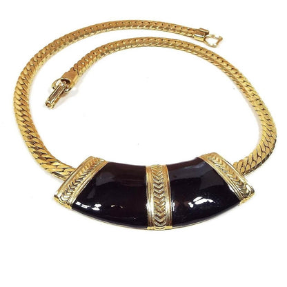 Front view of the retro vintage enameled bib necklace. The metal is gold tone in color. There is a wide herringbone chain that goes down to a curved elbow shaped bib area. The bib is black enameled with three gold tone stripes that have a chevron pattern on them. 
