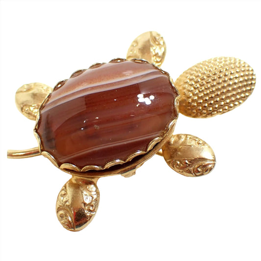 Angled top view of the retro vintage turtle brooch pin. The metal is gold tone in color. The head has a dot textured pattern. The legs have a stamped textured curly leaf like design. There is a fancy glass cab on the top made to look like a gemstone with orange and white stripes. The bezel on the glass cab is scalloped and you can see where the latch is on the side to open the shell up for the tiny compartment inside.