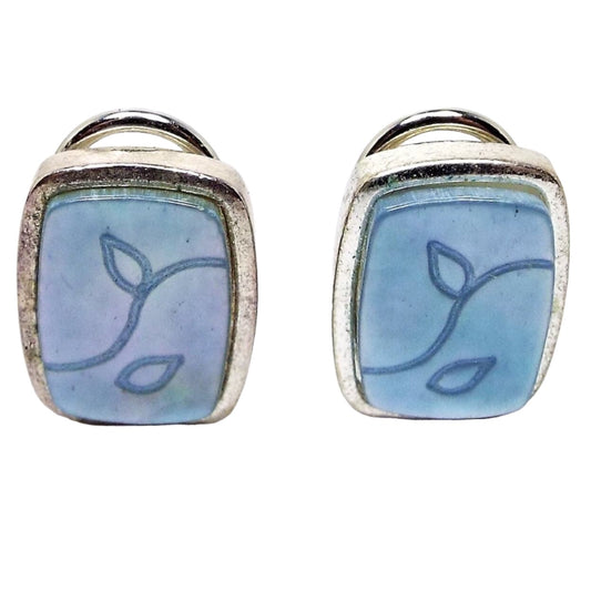 Front view of the retro vintage mother of pearl clip on earrings. The metal is silver tone in color and rounded rectangle in shape. On the front are light blue dyed mother of pearl shell cabs with a leaf design engraved on them.