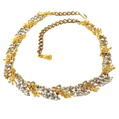 Front view of the Mid Century vintage BSK rhinestone link necklace. The metal is gold tone in color. Each link has a leaf like design and a cluster of six round clear rhinestones. There is a hook clasp at the end. The end chain is darker gold color than the rest of the necklace.