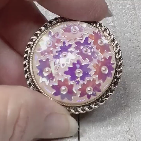 Video of the Mid Century vintage scarf clip made in West Germany. It's round and gold tone with a pink floral design. The video shows the iridescent areas of the scarf clip.