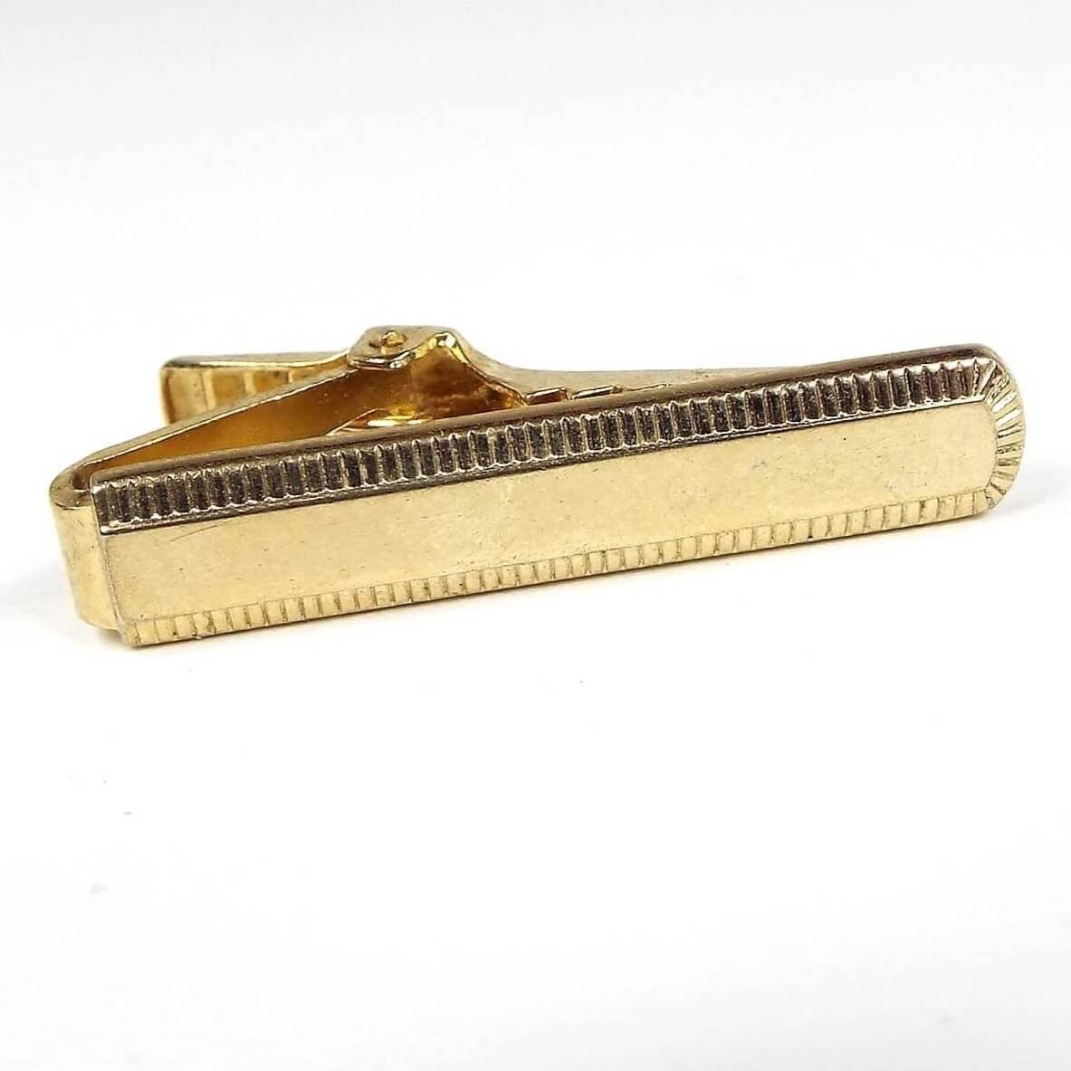 Front view of the Anson Mid Century vintage tie clip. The metal is gold tone in color. The end of the tie clip is rounded and there is a rectangle textured design around the edge of the tie clip.