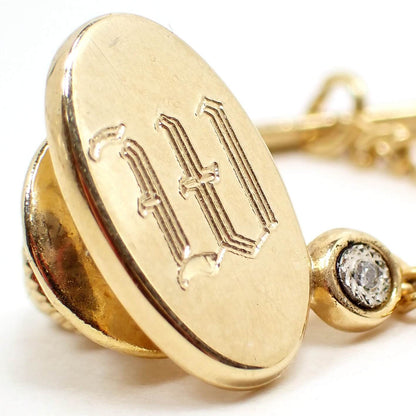 Enlarged angled front view of the retro vintage initial tie tack with diamond chip. The metal is gold tone in color and it is oval shaped. There is a fancy style block letter W engraved on the front. At the bottom right there is a small round area with faceted silver tone metal around a tiny diamond chip accent. 