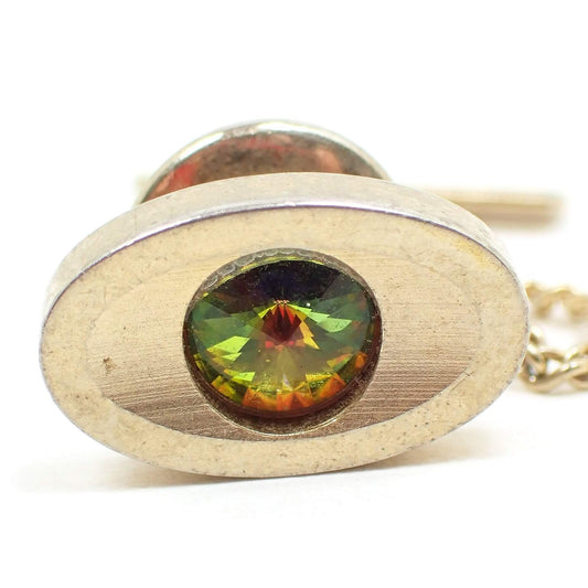 Enlarged front view of the retro vintage Sarah Coventry tie tack. It is oval in shape and has a matte textured front. The metal is gold tone in color. There is a rivoli rhinestone in the middle with a point going outwards that has flashes of green and orange color as you move around.