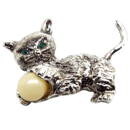 Front view of the Mid Century vintage cat brooch pin. the metal is silver in color. The cat has green rhinestone eyes and is holding a plastic ball that is yellow off white in color.