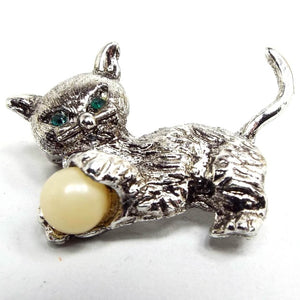 Front view of the Mid Century vintage cat brooch pin. the metal is silver in color. The cat has green rhinestone eyes and is holding a plastic ball that is yellow off white in color.