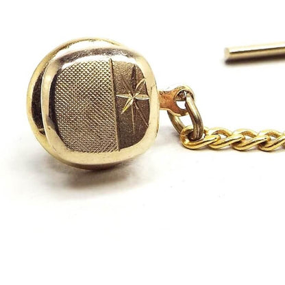 Enlarged front view of the Mid Century vintage Swank tie tack. It is gold tone in color and has a rounded square shape. There is matte brushed textured design on one side and an etched starburst design on the other. 